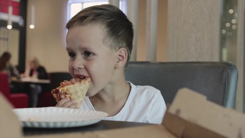 Portrait of a cute boy in a white t-shirt, who sits at the table in a pizzeria and appetizing eating a slice of sliced pizza, very tasty. Fast food
