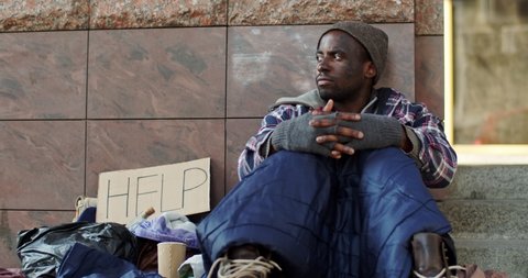 Young African American abondoned homeless guy in despair sitting on the ground with a table Help outside and being upset. Portrait.