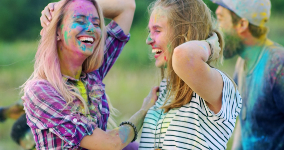 Two Cucasian cheerful young girls dancing and having fun among mixed-races friends at the holi festival. Outdoor.