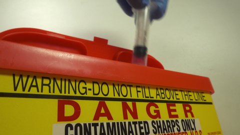 Northamptonshire, UK - November 5 2019 : A used sharps syringe being placed into a Sharps bin. The safe disposal of contaminated clinical waste for incineration. Biohazard needles syringes sharps.
