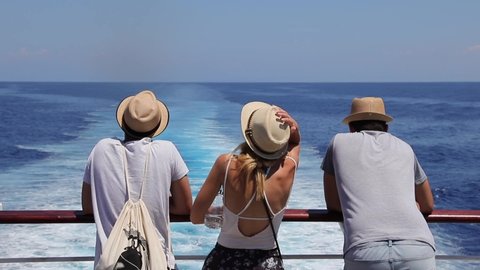Tourists with a straw hat stand on the deck of a cruise ship and look out over the ocean while the boat is sailing.