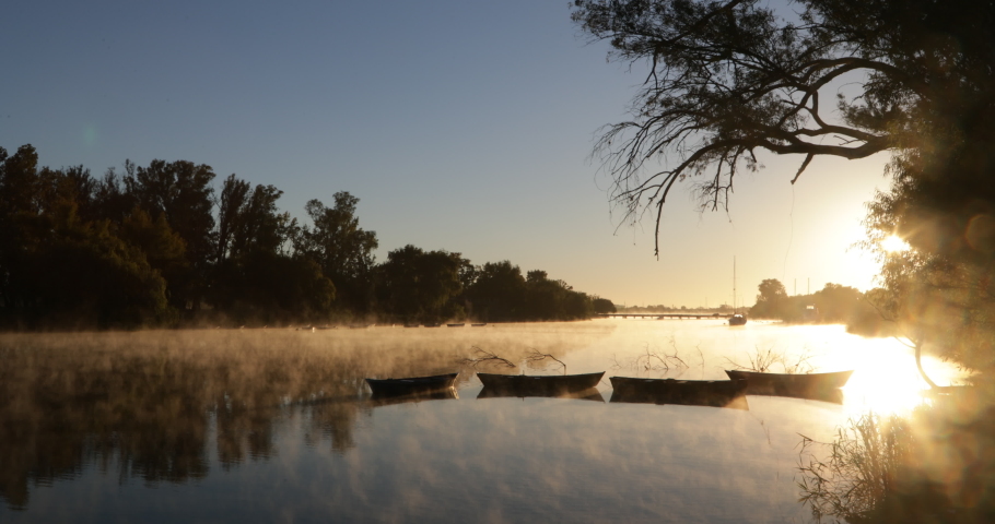 Golden foggy morning on river, movement of mist over water surface, silhouette of old rowing boats. Volume light and flares. Mysterious, calm scene. Rio Negro, Mercedes, Uruguay