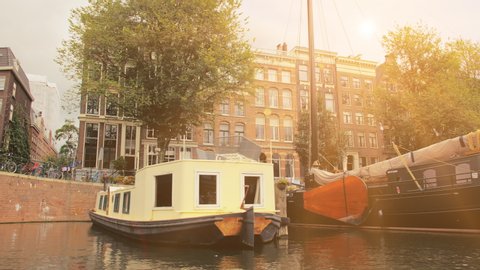 Amsterdam, Netherlands - September 2019: Lots of houseboats and ships moored in the streets. Facades of houses in the city center. The rays of sunset fall into the lens. View from the canal.