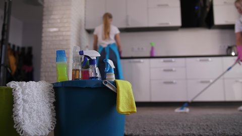 Two female janitors in aprons and gloves wiping kitchen surface and mopping, close-up on cleaning tools and detergents. Professional cleaners washing kitchen area during house clean up