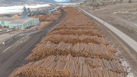 AERIAL: Flying over countless logs stacked into large piles next to a sawmill near a straight country highway running across the wintry rural landscape. Drone view of tree trunks and logging machinery