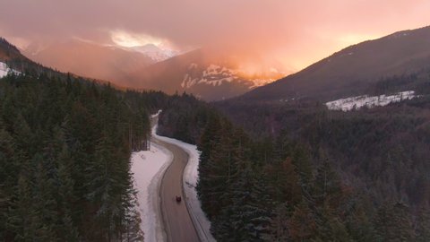 DRONE: Flying behind a car exploring the breathtaking sights of the Sea to Sky freeway at sunrise. Gorgeous winter evening sky illuminates the way for tourists on road trip driving through the woods.