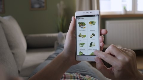 Woman Orders Food Home In An Online Store Using a Smartphone. Female Selects Vegetables in Grocery Online Store. Woman at Home Lying on Couch in Living Room Using Smartphone Buys in Internet Shop. 