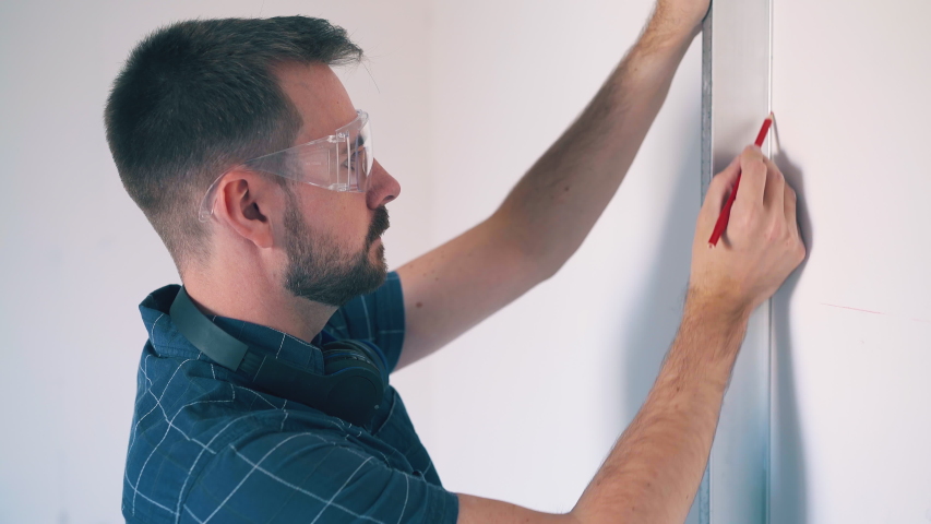 Tired man in blue shirt and goggles draws lines on white wall with modern level and pencil in spacious room closeup