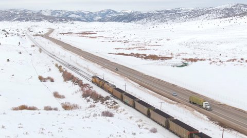 AERIAL: Cinematic shot of a train carrying coal across the country in winter. Cars drive along freeway running along the train tracks leading the locomotive transporting coal across the snowy valley.