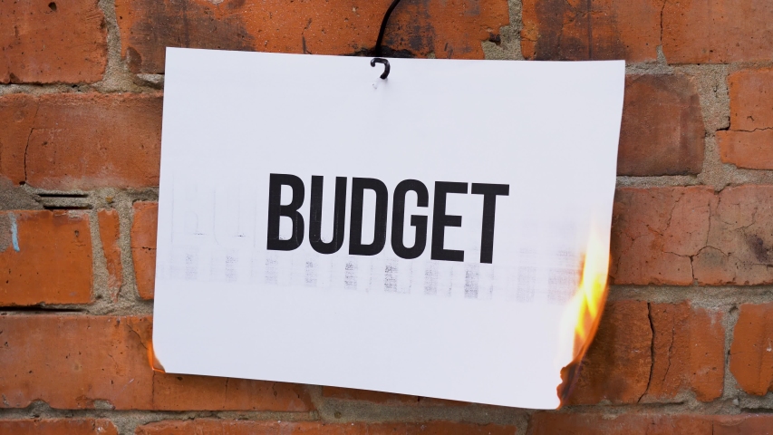 The inscription on a white A4 size sheet against the background of an orange brick wall. The paper with the black word Budget burns, smoke and turns into ashes. Business concept.
