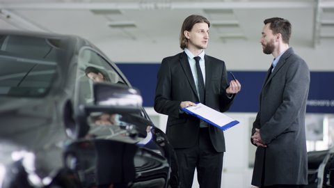 Professional car salesman is telling interested buyer about luxurious car in motor show while man is looking at auto and listening to dealer. They are discussing details of the sold car. Auto business
