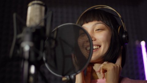 Closeup from below shot of young female Asian pop artist in earphones smiling while performing joyful song in microphone standing in booth in music recording studio