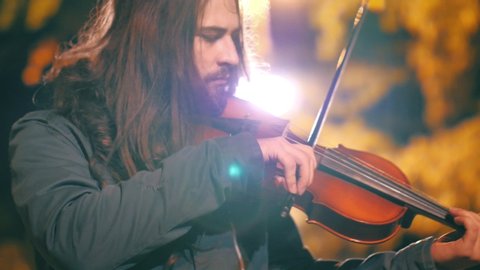 Handsome street musician rock violinist playing classical wooden violin, illuminated autumn city street in background. Closeup, shallow DOF, 4K UHD.	
