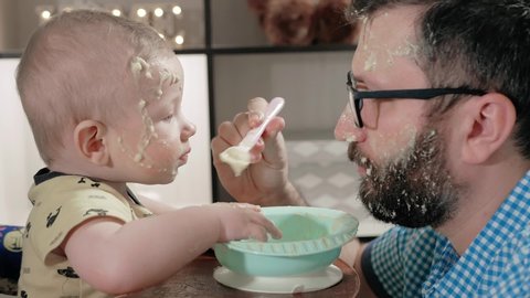 Father feeds baby from spoon. Dad and child have very dirty faces with porridge, baby behaves very messy, often puts his hand on plate and on face of parent. Close-up