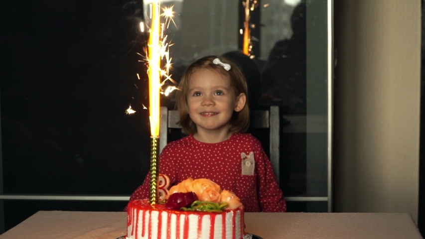 Portrait of little 3 year old smiling child girl celebrates birthday with festive cake with cream, candles, laughs and looks with delight at sparkling fireworks. Happy holiday concept
