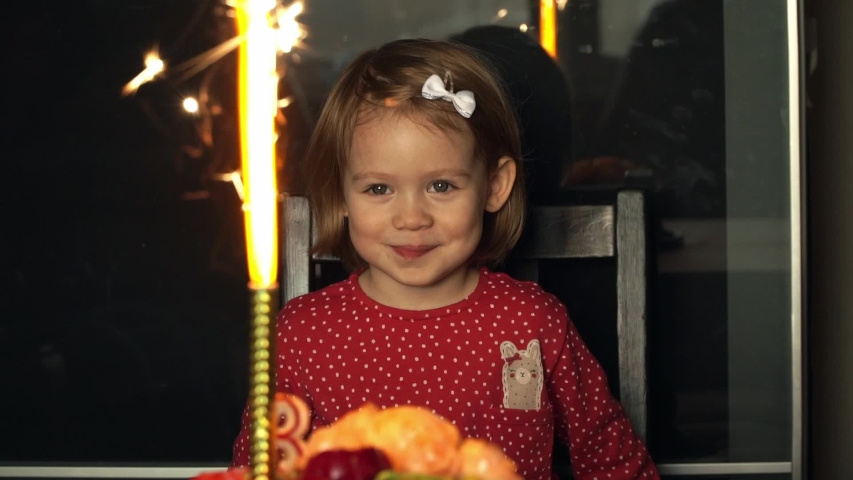 Close up of portrait of little smiling 3 year old child girl celebrates birthday with festive cake with cream, candles, laughs and looks with delight at sparkling fireworks. Happy holiday concept