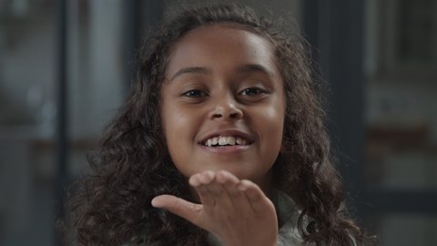 Portrait of charming elementary age african american girl with long curly hair blowing kisses, looking playfully with cheerful radiant toothy smile, expressing positivity and happiness.