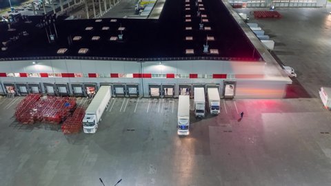 Aerial hyper lapse (motion time lapse), top view of the large logistics park with warehouse, loading hub with many semi-trailers trucks standing at the ramps for load/unload goods at night