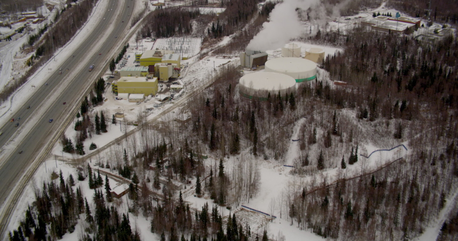 Aerial helicopter shot, tilt down to see steam rising from power plant, cars pass on highway, snow in background, drone footage