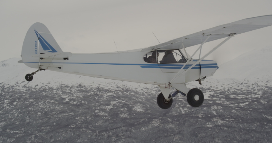 Aerial helicopter shot, small blue and white airplane banking left across vast Alaskan mountain valley, drone footage