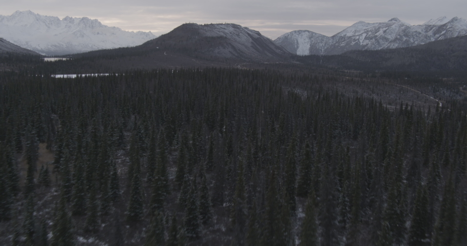 Aerial helicopter shot above winter valley with dark evergreens, low over partially frozen lake towards mountains, drone footage