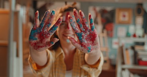 Portrait of the young cheerful Caucasian pretty girl painter smiling to the camera over the hands palms in red ad blue paints which she showing to the camera. Close up.