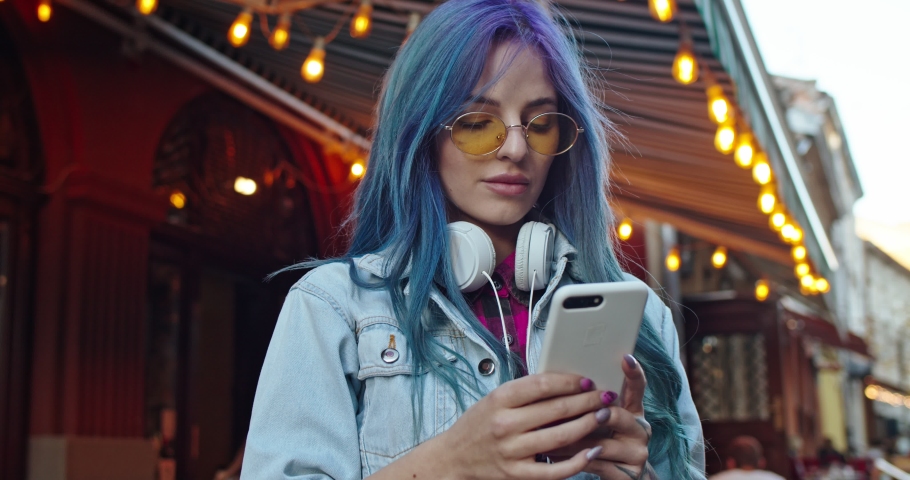 Portrait shot of the Caucasian stylish beautiful young hipster girl with blue hair and in sunglasses typing and tapping on the phone, texting a message, then smiling cheerfully to the camera. Outdoor.