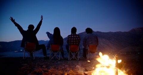 Silhouettes of four friends sitting in chairs near bonfire on top of mountains, watching the sun set and chatting. enjoying their trip - friendship, youth concept 4k footage