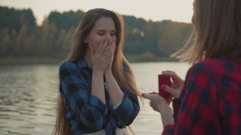 Woman proposing to her beloved sweetheart with engagement ring at beautiful lake. Excited adorable girl accepting her girlfriend marriage proposal hugging in nature outdoors.