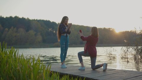 Young happy woman proposing to her excited girlfriend outdoor at sunset. Affectionate girl accepting proposal hugging her darling kissing enjoying moment of love. Marriage concept.
