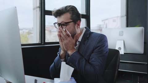 Overworked young business man suffering bad feeling or cold coughing at working desk. Poor ill employee sneezing allergy using paper tissues sitting in the office.