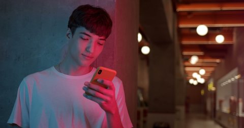 Young Stylish Millenial Boy in White T-Shirt Using his Smartphone for Chatting, Typing Messages while Standing at Futuristic Neon Club Lights Background. Close Up Shot.