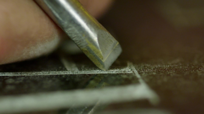Macro view of process of manual engraving letters on surface of dark granite block as part of headstone, some worker's hand is using metallic tool for this purpose.