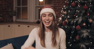Smiling Young Pretty Girl Wearing Santas Hat Sitting near Christmas Tree Waving Hi Holding and Giving Red Present Box Greeting Laughing Blowing a Kiss Making a Video Call or Video Message. Concept of