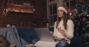 Happy Caucasian Girl Wearing Christamas Hat Sitting on the Sofa at Cosy Home Background near Christmas Tree. Woman Making a Video Call Using Her Laptop Talking Showing a Tree. Concept of Holidays and