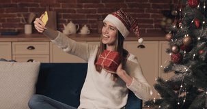 Happy Caucasian Young Pretty Girl Wearing Christamas Hat Sitting on the Sofa Holding and Shaking Present Box near Christmas Tree Making a Video Call or Video Message. Concept of Holidays and New Year