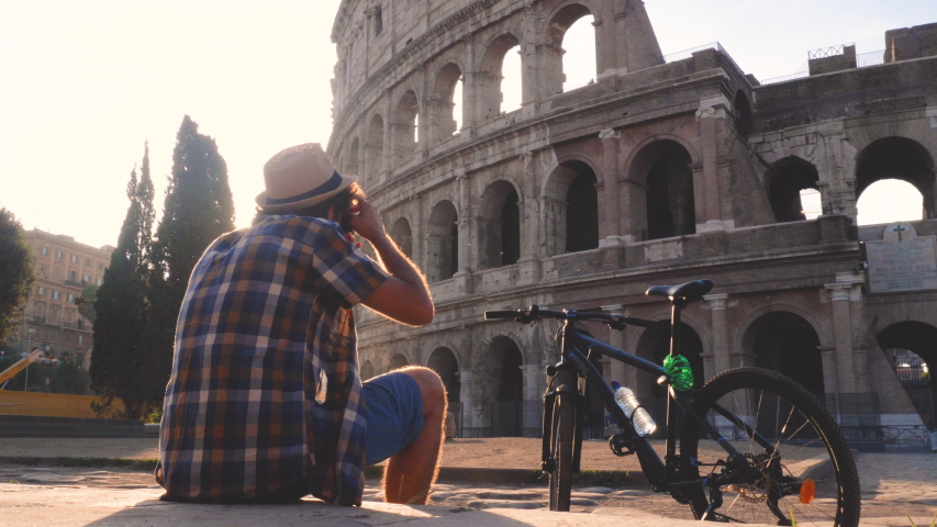 Happy young man tourist wearing shirt and hat with bike sitting and taking pictures with vintage camera at colosseum in Rome, Italy at sunrise.