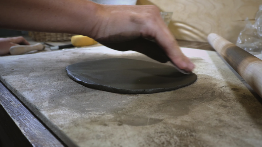 Closeup of female potter's hands prepares dark brown clay blank. Woman removes roughness from a clay blak with a blade. Potter's workplace