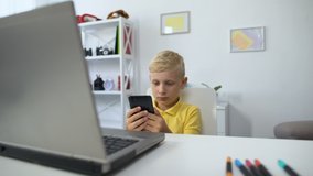 Little male kid sitting in front of laptop and watching video on smartphone.