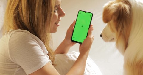 GREEN SCREEN tracking dot. Portrait of Pretty girl student is using smartphone shows screen and laughing lying on bed at home with cute pet dog border collie , animal is enjoying care and love.