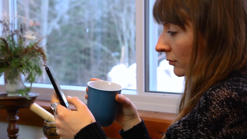 Young woman with wooly grey shirt is laughing out loud while scrolling on her phone at home. Fixed Angle Closeup