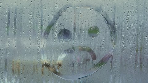 Woman draws a finger on the cold condensation window with water drops sad smile. Conceptual design for symbol of failure, longing, sadness.