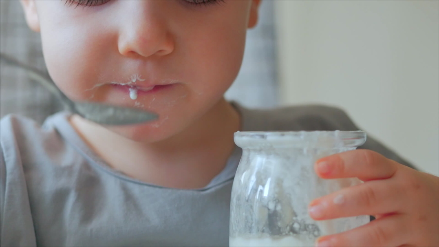 Close-up of a baby sitting at the table and eating baby food with a spoon on its own. A 2-3 year old boy eagerly eats milk yogurt or kefir, sour cream, baby food. 4 K