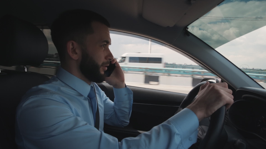 Handsome businessman talking on mobile phone and watching at road while driving car. American young man having business talk, holding steering wheel in hand sitting in black interior of automobile | Shutterstock HD Video #1040697689