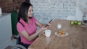 Happy disabled woman having video call using phone at table in kitchen interior. Portrait of american paraplegic female talking with smile, watching in camera holding smartphone in hand, sitting in