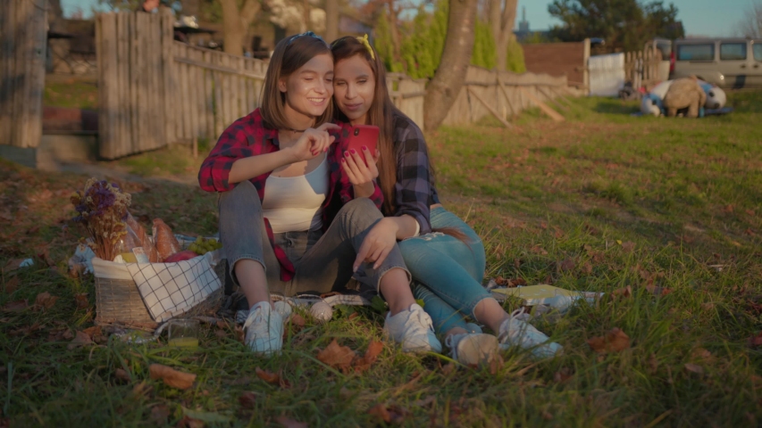 Pretty positive girlfriends hugging relaxing on the grass laughing of fun internet smartphone content enjoying autumn evening in the countryside.