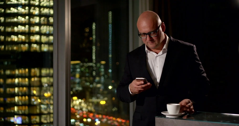 a middle-aged man in glasses and a suit checks his phone and drinks coffee by the big window. outside the window is dark and the lights of buildings glow. blurred lights of cars move and flicker