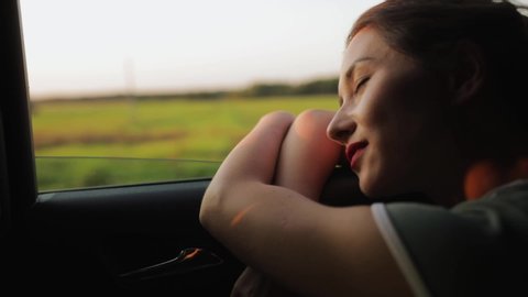 Cinematic inspirational video of young woman travelling by car or camper van, opens window to breathe fresh air of countryside, moves hand in wind. Sings melody of song, summertime vacation vibes