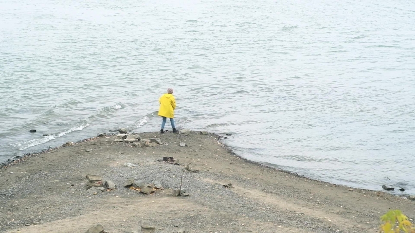 young girl in knitted cap and yellow raincoat walks along rocky Bank of river on an autumn cloudy day. A woman throws stones into the water and enjoys the peace and quiet in nature. weekend in nature.