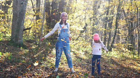 Happy family: young mother with little cute daughter is having fun, laughing and throwing yellow leaves at autumn forest together. Leaves is falling in slow motion. Fall season concept.